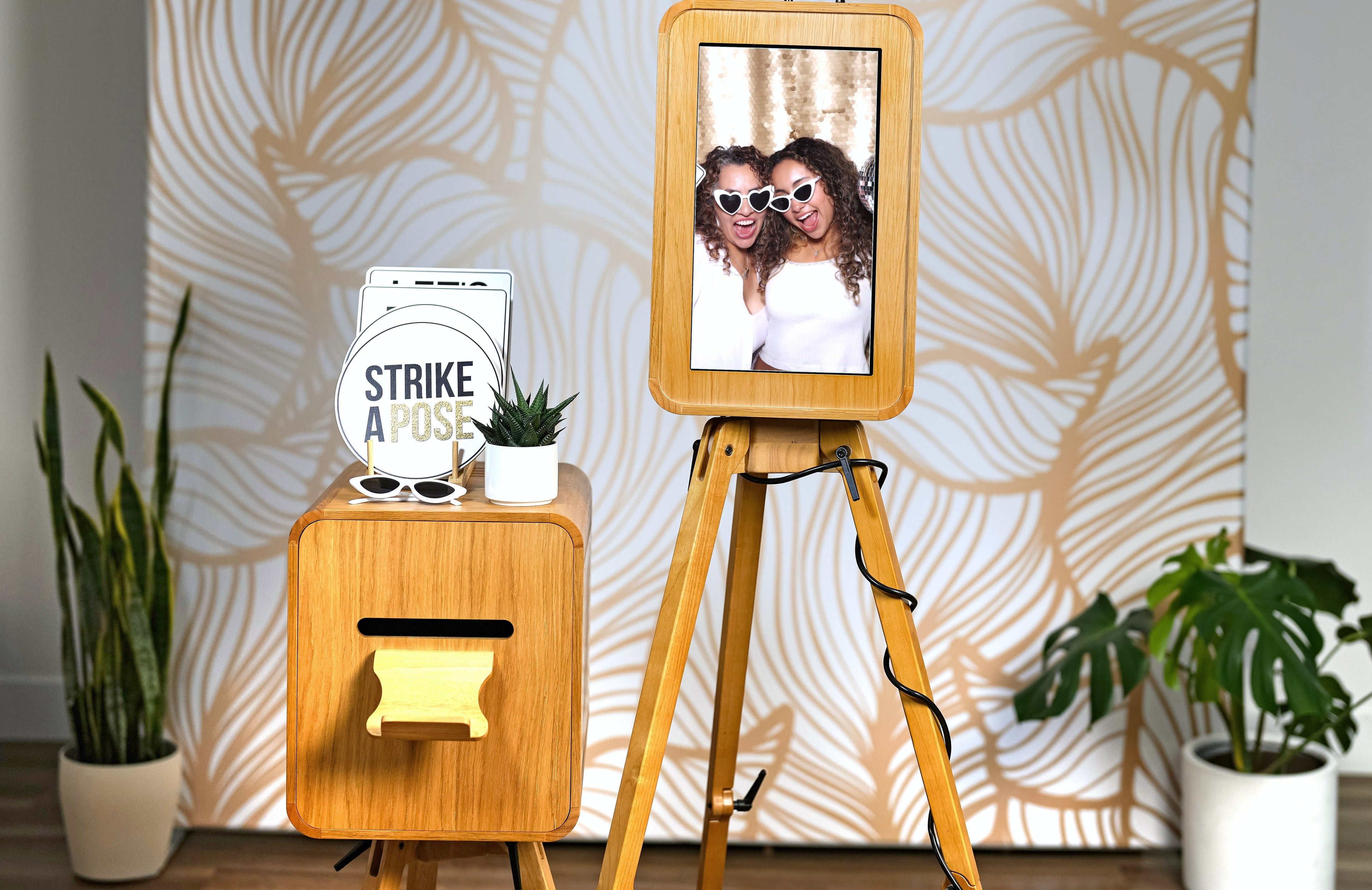 The Shutter Bar photo booth on its wooden tripod against a botanical backdrop, its screen showing guests posing with props.